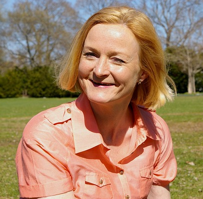 a middle aged woman with strawberry blond hair, sitting on grass and smiling at the camera